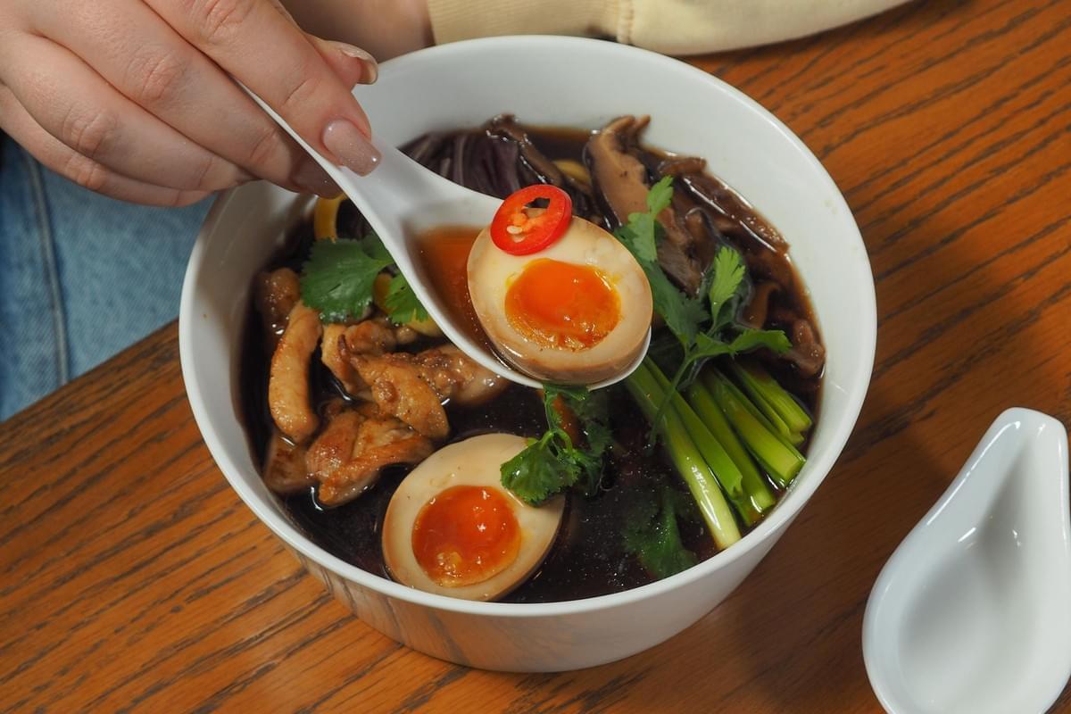 A person uses a white soup spoon to scoop a soft-boiled ramen egg topped with a slice of red chili pepper from a bowl of hot ramen. The bowl is filled with dark savory broth, chicken, mushrooms, fresh cilantro, and green vegetables, set on a wooden table.