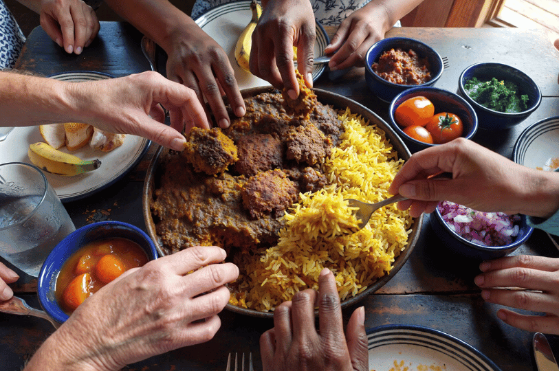 A communal dining scene showing multiple hands sharing a large platter of bobotie and yellow rice, surrounded by small bowls of sambals and fruit.