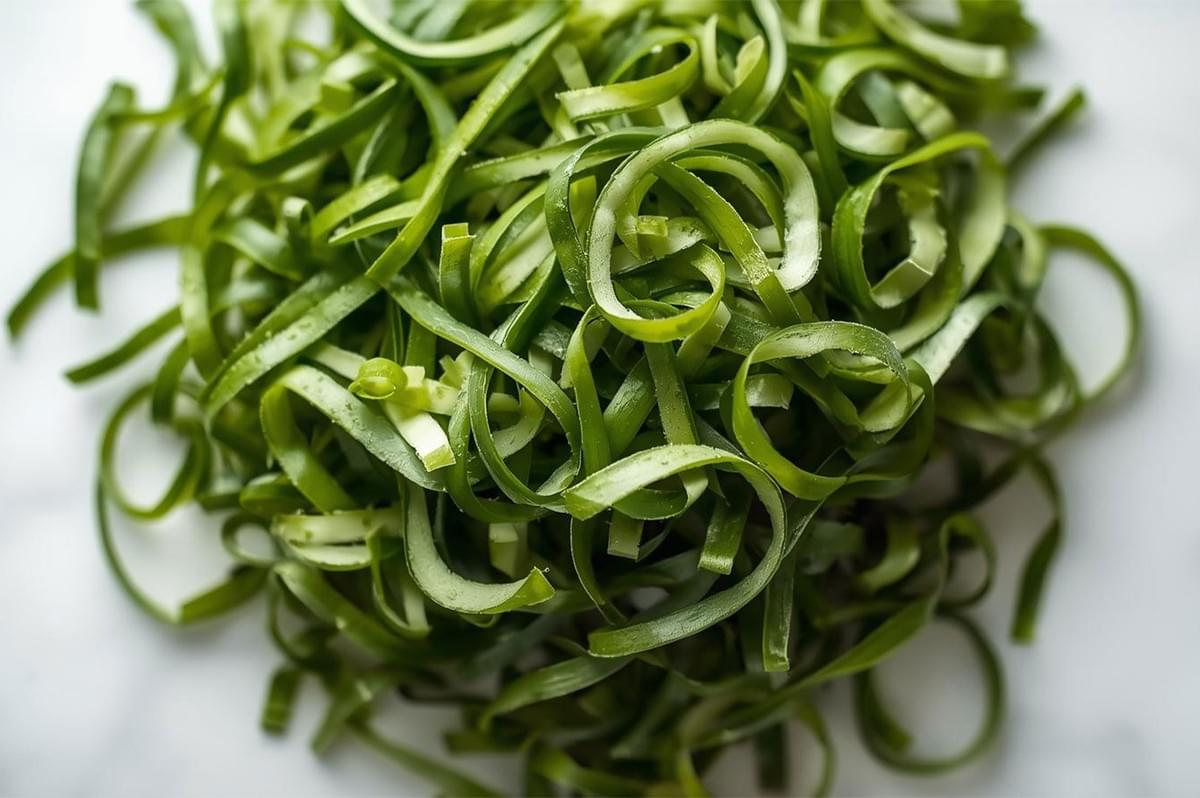 A detailed, close-up overhead shot of a mound of thinly sliced, curly dark green herbs (likely basil, mint, or scallions), demonstrating the chiffonade cutting style.