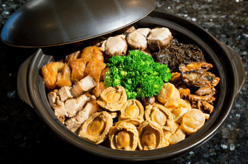 A top-down view of a traditional clay pot Pen Cai filled with premium abalone, sea cucumber, roast pork, and broccoli, partially covered with a lid on a dark marble surface.