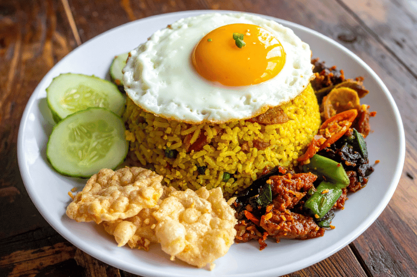 A bright, colorful plate of yellow-tinted Nasi Goreng topped with a sunny-side-up egg, served with a side of spicy beef rendang or stir-fried meat, cucumbers, and crispy crackers.