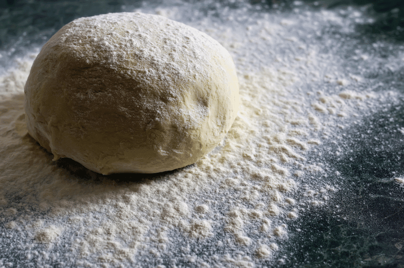 A large, rounded ball of dough resting on a dark countertop heavily dusted with flour, showing a smooth surface indicating early gluten structure.