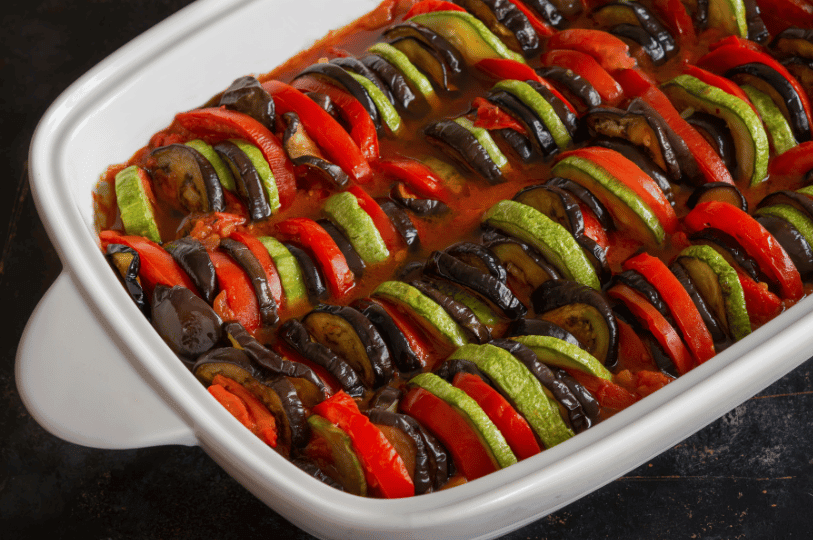 A white rectangular baking dish filled with a classic Provencal Ratatouille, featuring tightly packed, alternating slices of vibrant red tomatoes, green zucchini, and purple eggplant baked in a rich tomato sauce.