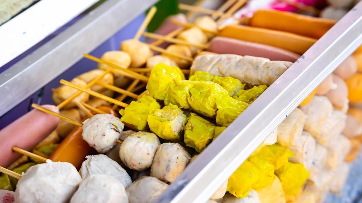 This image shows an angled, close-up view of various street food items, including meatballs, sausages, and yellow dumplings, all threaded onto wooden skewers.