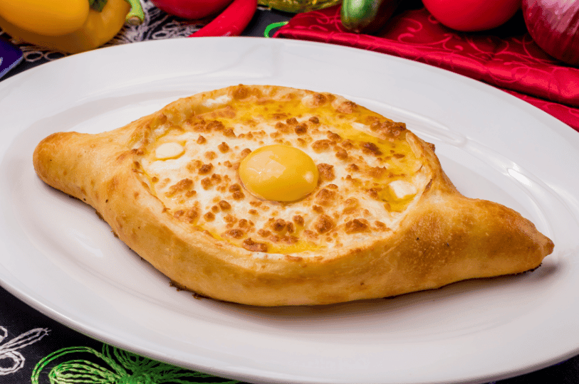 A close-up, side-angle shot of a freshly baked Georgian Khachapuri. The crust is perfectly tanned, and the center is filled with a mixture of molten white cheese and a glossy yellow egg yolk, served on a white ceramic plate with colorful peppers in the background.