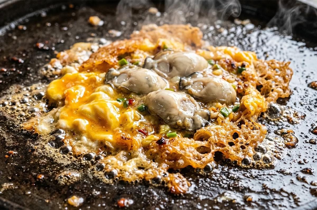Sizzling oyster omelet being cooked on a flat griddle, showing crispy eggs, green onions, and fresh oysters.