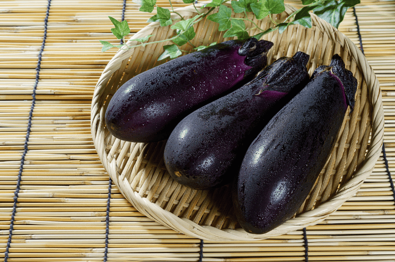 Three glossy, dark purple Summer eggplants with water droplets on their skin, resting in a woven bamboo basket on a traditional tatami mat.