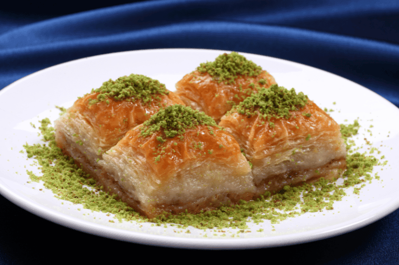 Four square pieces of baklava arranged on a white plate, topped with vibrant green pistachio crumbles and set against a deep blue fabric background.