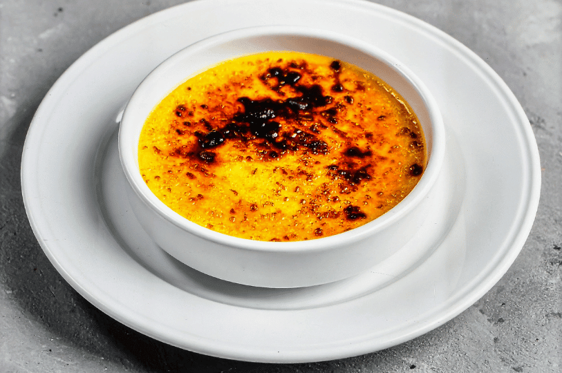 High-angle shot of a single crème brûlée on a white plate, highlighting a rustic, unevenly caramelized sugar crust with dark charred spots for a bittersweet flavor.