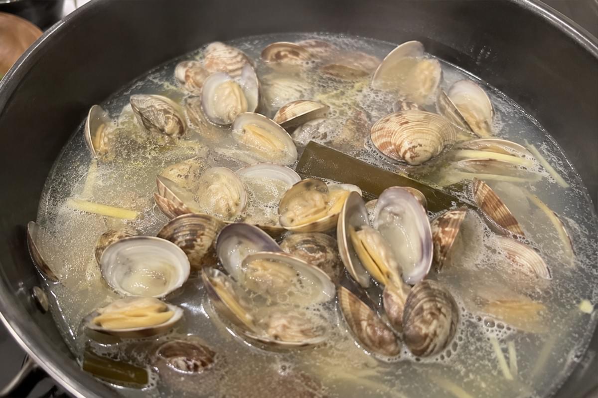 A high-angle, close-up shot shows numerous clams cooking inside a large black metal pot filled with a simmering, bubbling broth. Nearly all of the variegated brown and white clam shells have opened up, exposing the cooked meat inside. Strips of julienned yellow ginger are scattered throughout the liquid, and a single thick, dark green piece of kombu kelp sits submerged in the middle of the pot amid the gentle foam.
