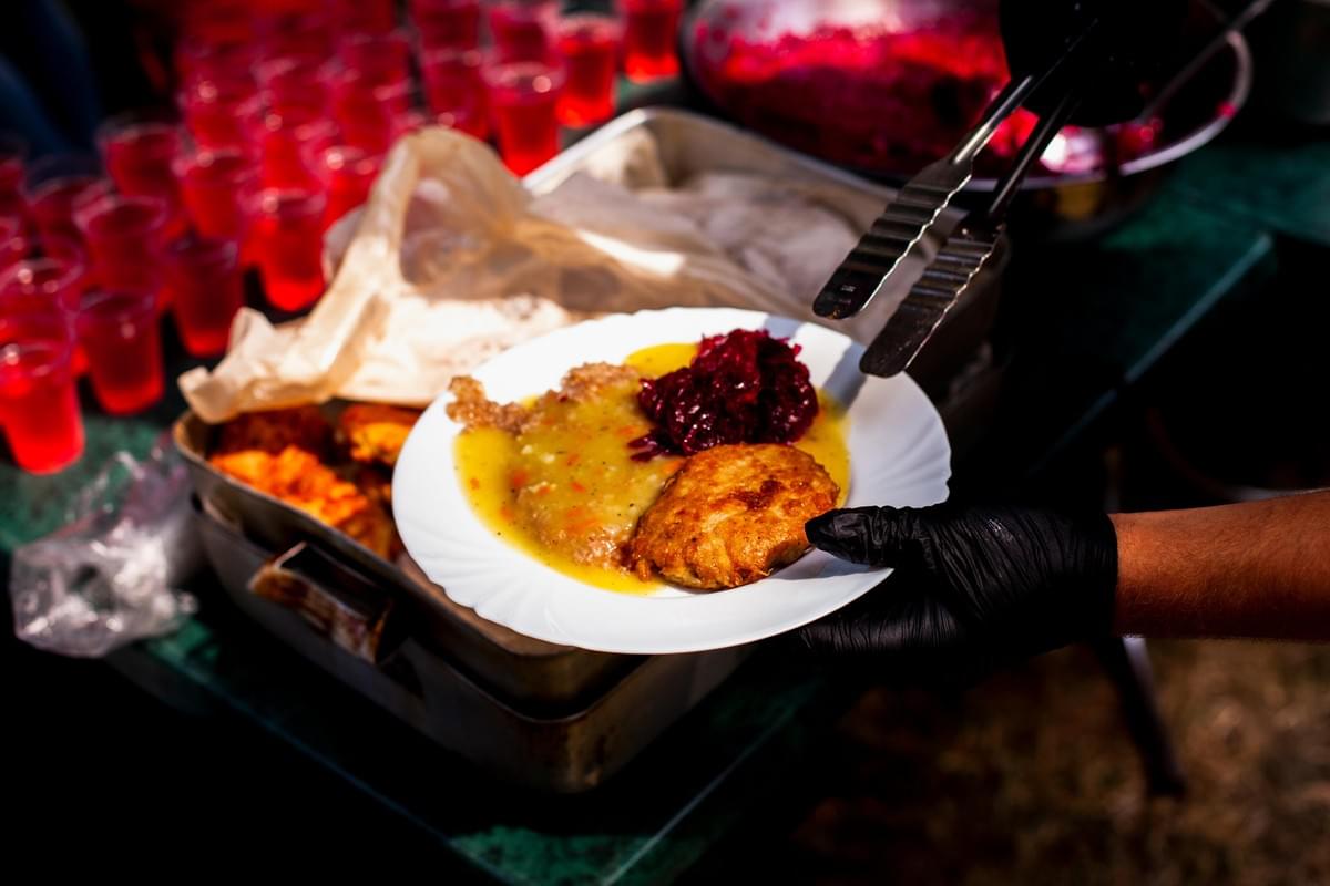 In the image, a person wearing a black glove holds a white plate filled with a golden-brown meat cutlet, gravy, and a dark red beet salad. Metal tongs hover above the plate at an outdoor buffet setup, with numerous small cups of bright red drinks visible in the background.