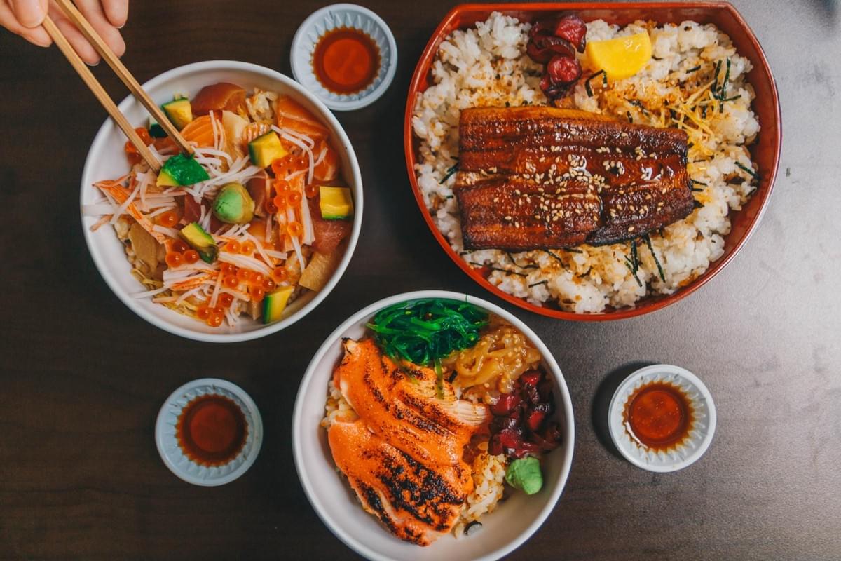 A top-down view shows three Japanese rice bowls, including a chirashi bowl with salmon and avocado, a grilled unagi bowl, and a seared salmon bowl, accompanied by small saucers of soy sauce. A hand holds a pair of wooden chopsticks over the chirashi bowl, dipping into the toppings.