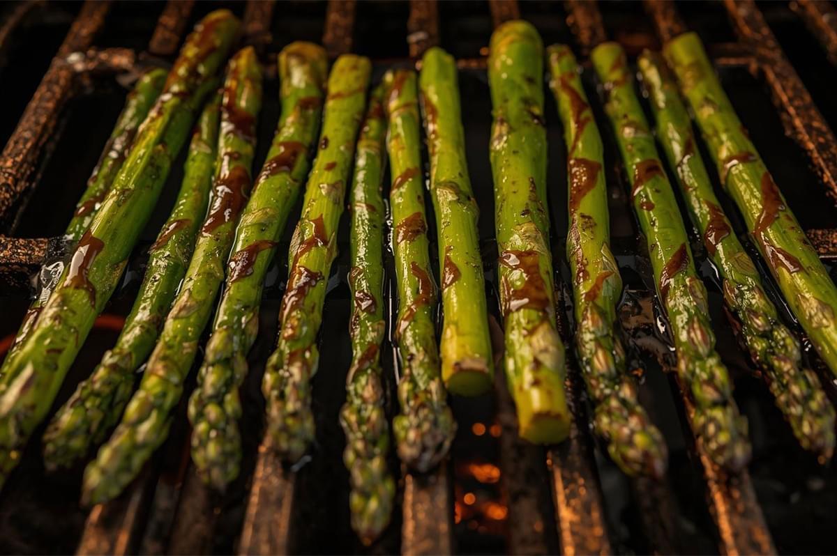 A close-up shot of several asparagus spears lying directly on a hot grill grate, showing smoky char marks and a light glaze, ready to be served.
