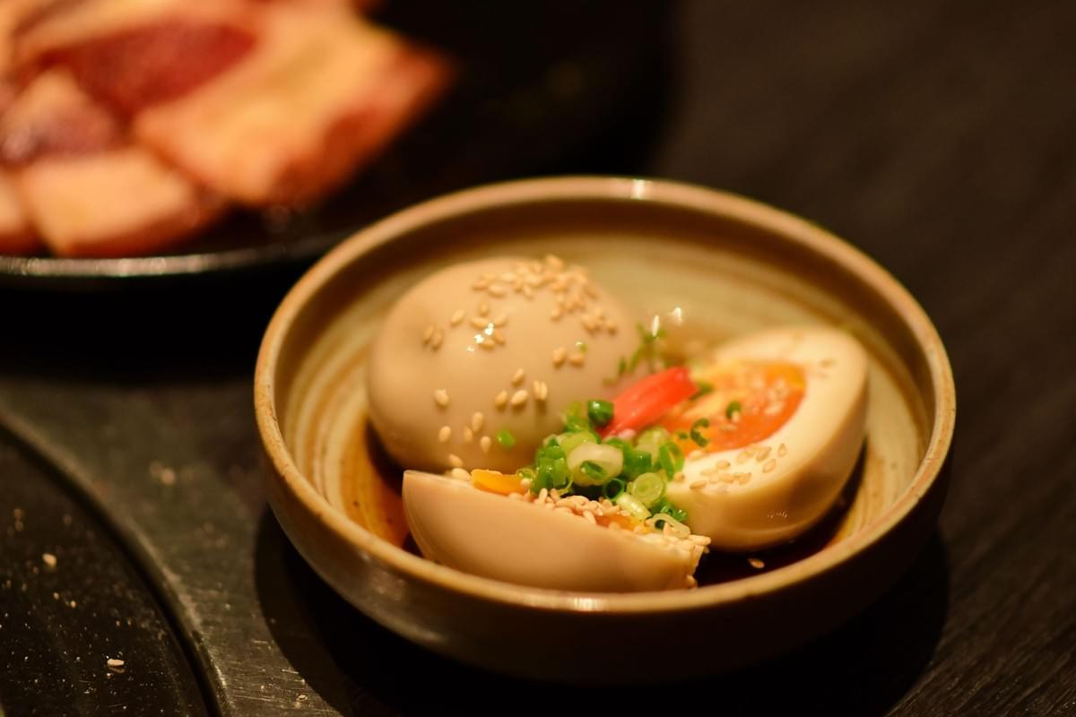 A small ceramic bowl holds soft-boiled, soy-marinated eggs topped with sesame seeds, chopped green onions, and a red chili slice. The dish rests on a dark tabletop alongside another plate of food in warm, ambient lighting.