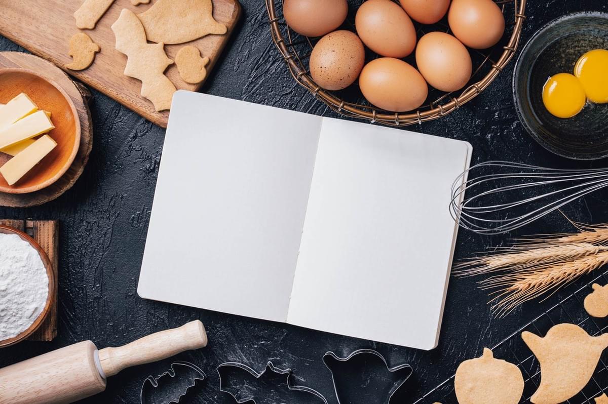 Flatlay of baking ingredients with an open blank recipe notebook, surrounded by eggs, flour, butter, cookie cutters, and kitchen utensils.