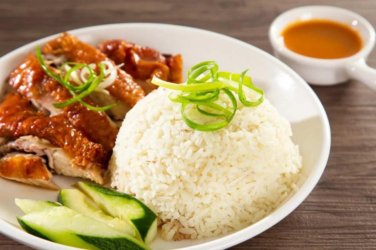 A serving of Singapore Chicken Rice with succulent chicken, aromatic rice, and fresh vegetables on a white plate.