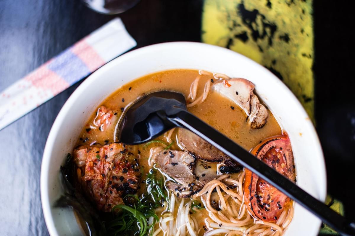 A top-down, close-up view of a bowl of ramen filled with broth, noodles, chashu pork, a slice of grilled tomato, green onions, bean sprouts, and kimchi topped with black sesame seeds. A black soup spoon rests inside the white bowl, which sits on a dark surface next to a paper-wrapped pair of chopsticks.