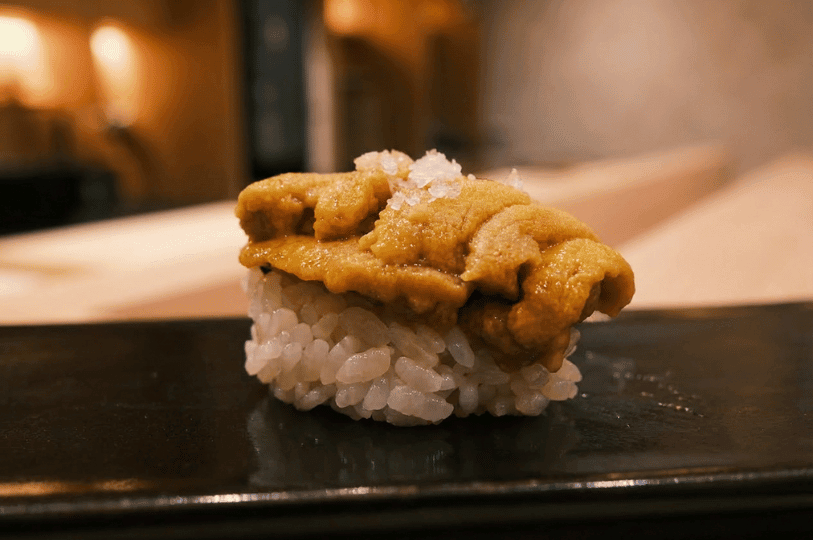 A delicate piece of seasonal uni nigiri served on a rectangular black ceramic plate resting on a light wooden sushi bar at Sushi Masa by Ki-setsu.