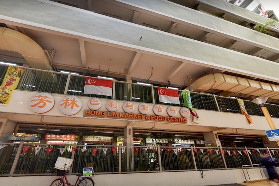 This low-angle shot features the exterior of the Hong Lim Market & Food Centre, adorned with Singaporean flags and circular signage. Various food stalls and a parked bicycle are visible on the ground level beneath the building's concrete tiers.