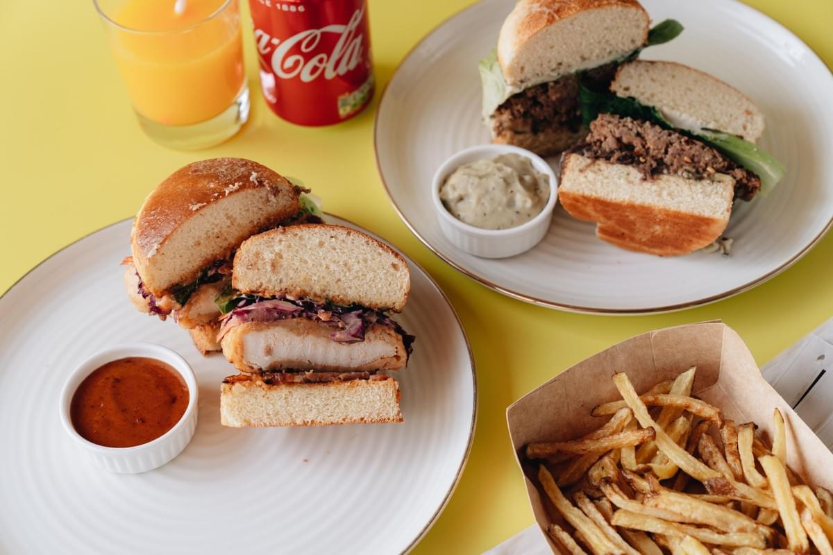 A bright yellow table is set with two plates of halved gourmet burgers, small ramekins of dipping sauce, and a container of golden french fries. A glass of orange juice and a red Coca-Cola can sit nearby, completing the vibrant meal spread.