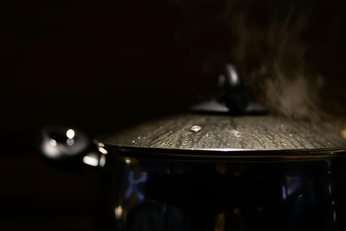 Condensation clings to the glass lid of a dark, metallic pot as steam escapes from the side. The shot uses a shallow depth of field, blurring the handles and the rising vapor against a black background.