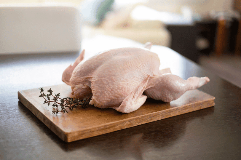 A raw whole chicken resting on a wooden cutting board next to a sprig of fresh thyme.