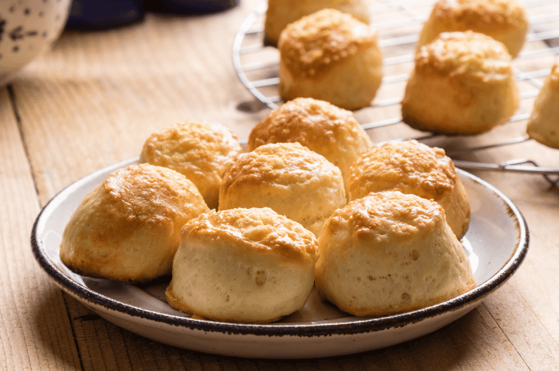 A white ceramic plate filled with golden-brown, round plain scones, with more scones cooling on a wire rack in the background on a wooden table.