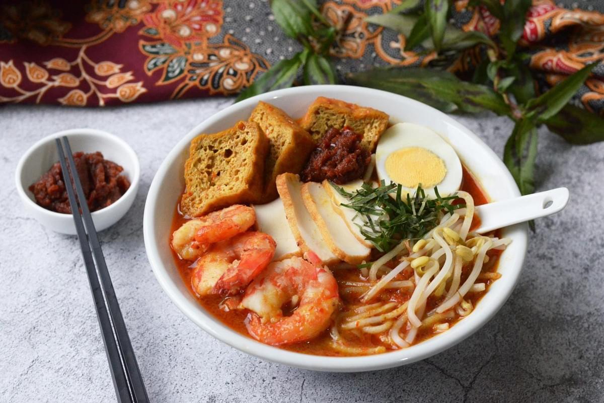 A vibrant bowl of Singapore Laksa is filled with thick rice noodles, prawns, tofu puffs, and a hard-boiled egg in a rich coconut curry broth. Beside the main dish sits a small bowl of spicy sambal paste, all set against a textured grey background and traditional batik fabric.