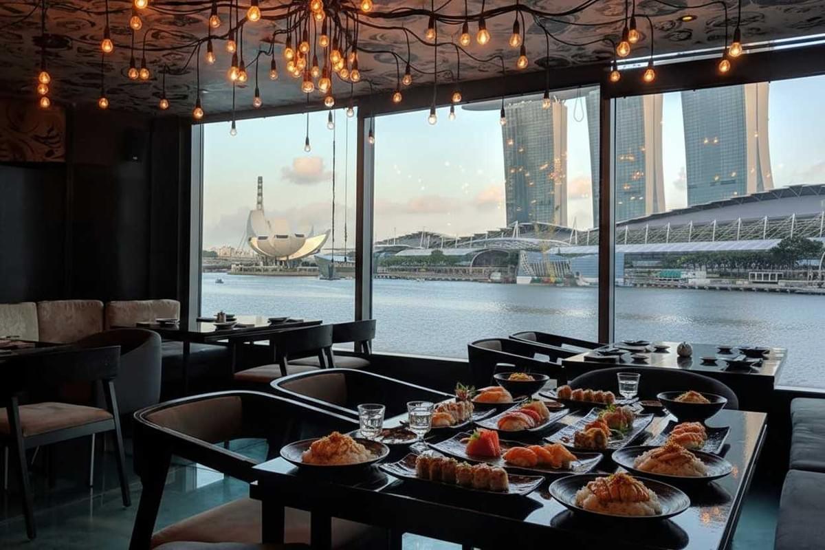 This image features a spread of sushi and Japanese dishes on a dark table inside a restaurant with warm, industrial-style lighting. Large windows offer a stunning waterfront view of the Singapore skyline, including the ArtScience Museum and Marina Bay Sands.