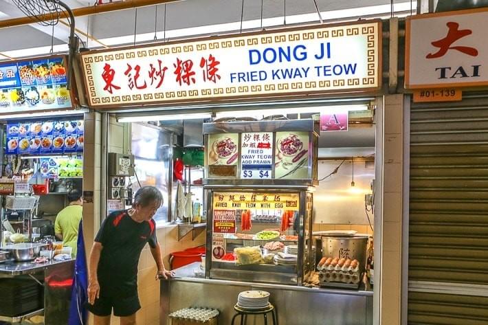 The image depicts a food stall named Dong Ji Fried Kway Teow, featuring a lighted sign with traditional Chinese characters. A man in a black t-shirt stands in front of the stall, which displays various ingredients and pricing for its noodle dishes.