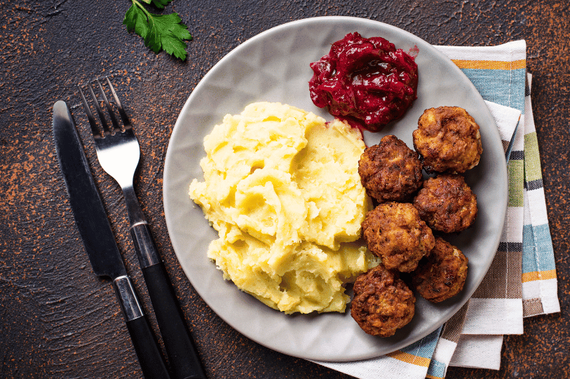 An overhead shot of a classic Swedish meal on a grey plate, showing the contrast between the crispy, dark exterior of the meatballs, the pale yellow mashed potatoes, and the vibrant red lingonberry sauce, with a fork and knife to the side.