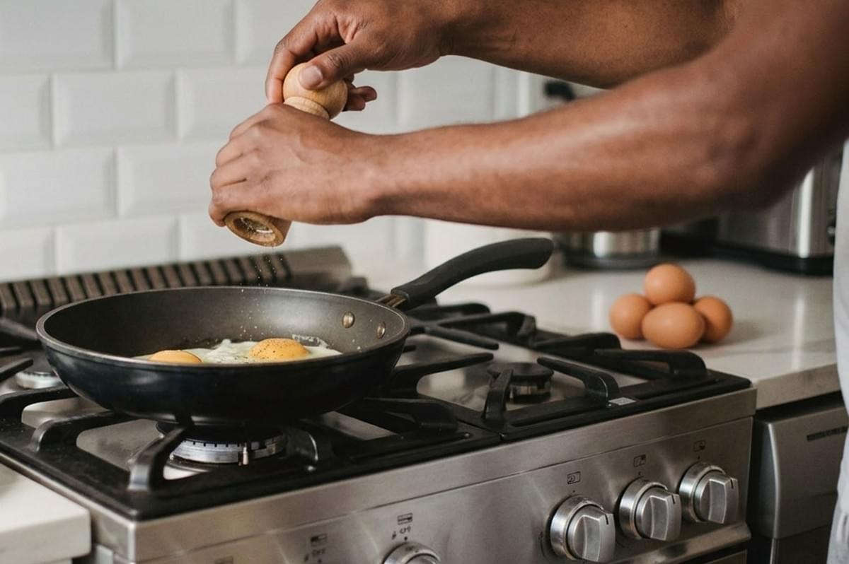 Person seasoning eggs in frying pan on gas stove with pepper grinder