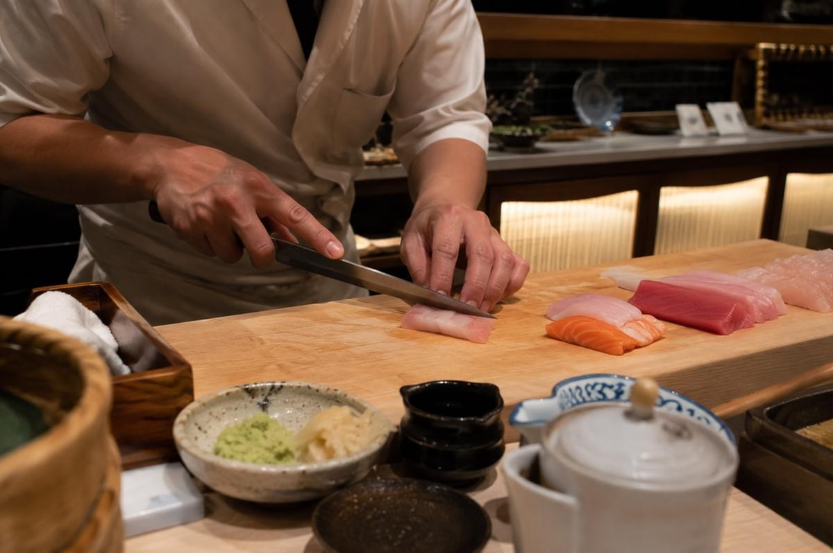 A close-up, eye-level shot of a sushi chef in a crisp white jacket slicing a piece of pale pink fish on a wooden cutting board with a sharp, traditional yanagiba knife, surrounded by freshly prepared slices of salmon and tuna, along with ceramic dishes of freshly grated wasabi, pickled ginger, soy sauce, and a white soy pot in the foreground.