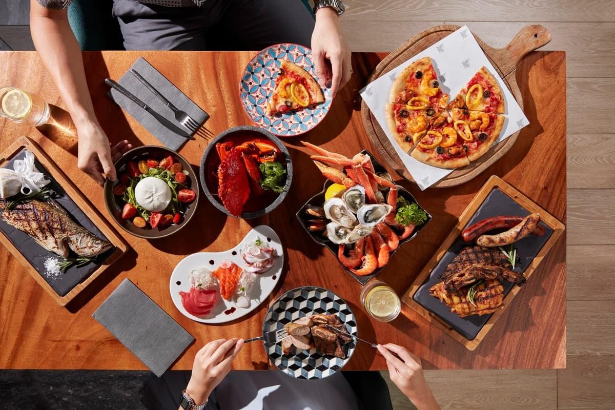An overhead view captures a lavish buffet spread arranged on a warm wooden table, where people are gathered to share a meal. The diverse feast includes a seafood platter with crab legs and oysters, a whole grilled fish, a burrata salad, a plate of fresh sashimi, grilled meats, and a seafood pizza.