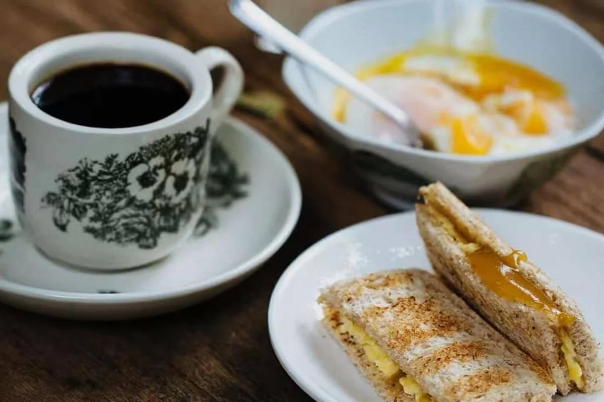 A cup of black coffee in a floral cup sits next to a plate with buttered kaya toast. A bowl of soft-boiled eggs is in the background on a wooden table.