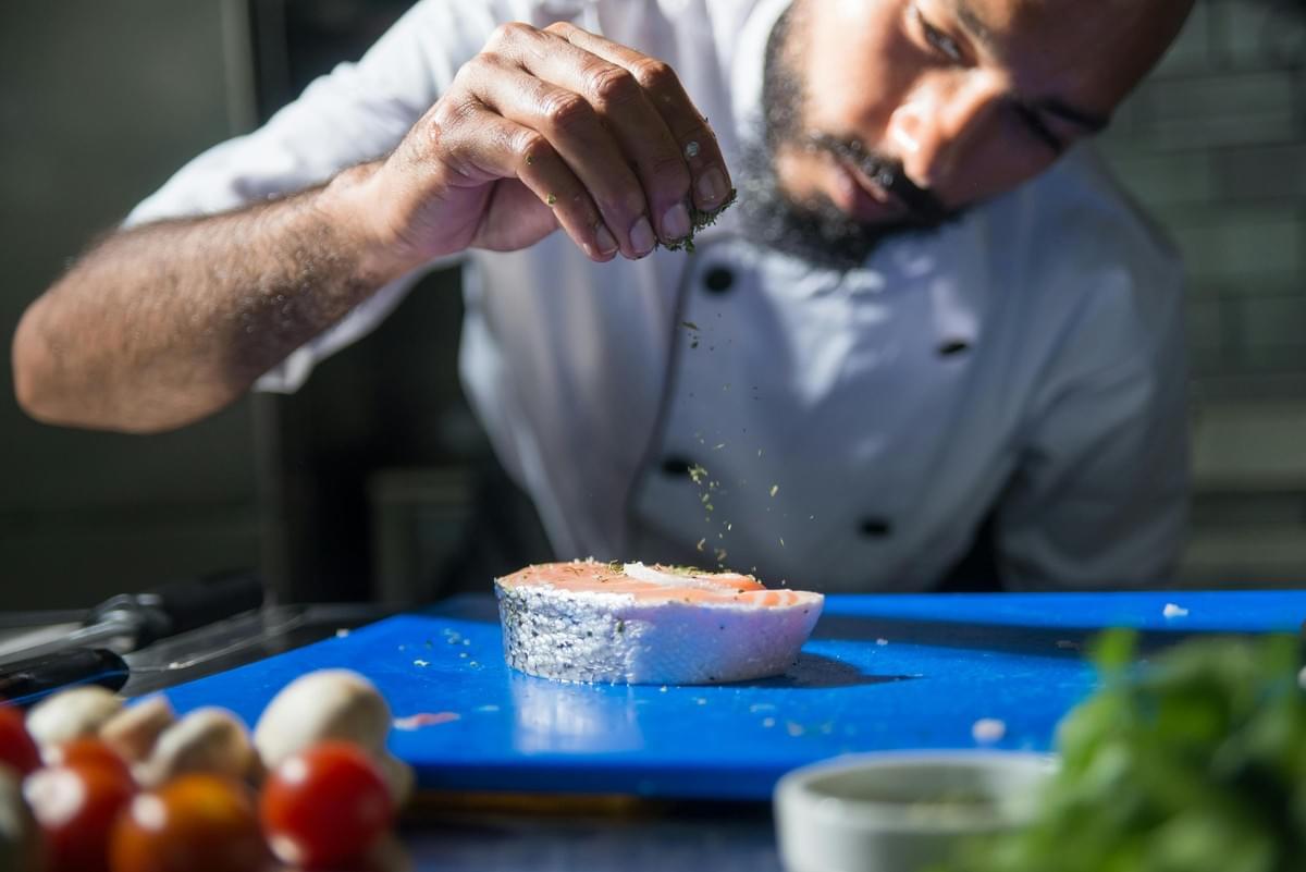  This image captures a chef, wearing a white uniform and sporting a beard, intently seasoning a thick cut of salmon steak with dried herbs by sprinkling them from his hand.