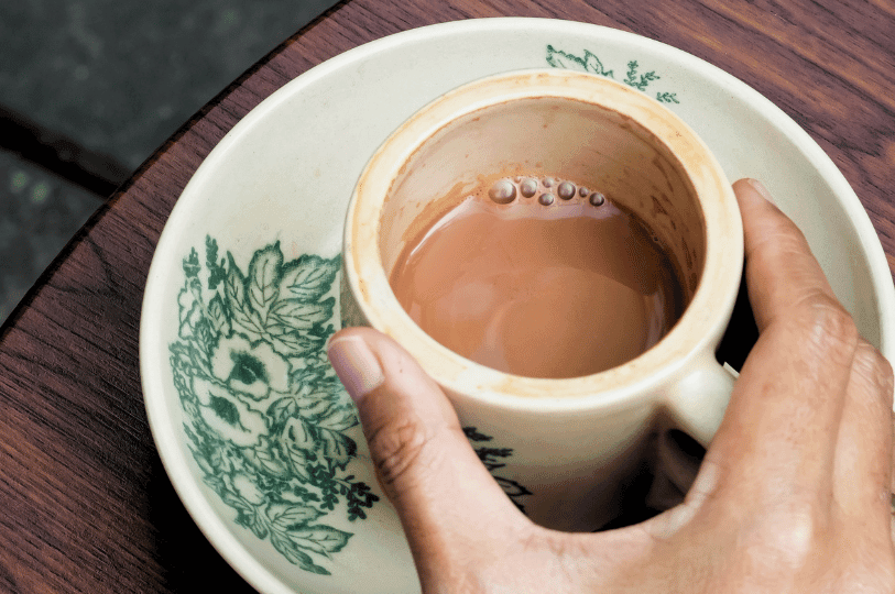 A top-down view of a person's hand holding a traditional floral ceramic cup filled with light brown, milky Singapore kopi. The coffee has a slight foam with bubbles on the surface and rests on a matching saucer.