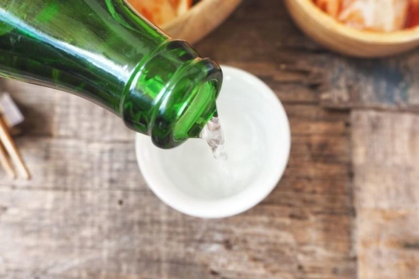 A close-up shot shows a clear, colorless liquid being poured from a green glass bottle into a small white ceramic cup. The drink is served on a rustic wooden table, with blurry wooden bowls containing food visible in the background.