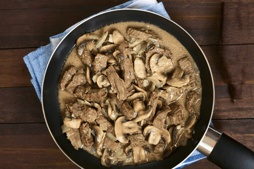 A top-down view of a frying pan resting on a blue kitchen towel, filled with simmering beef strips, sliced mushrooms, and onions in a savory brown sauce for Russian Beef Stroganoff.