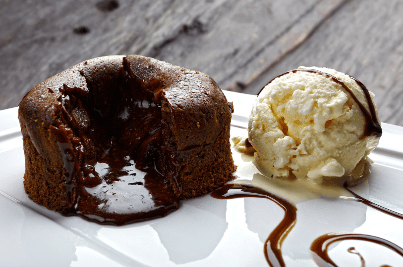 A close-up shot of a warm chocolate lava cake next to a melting scoop of vanilla ice cream. The liquid chocolate center has pooled onto the white plate, illustrating the ideal "molten" consistency achieved through precise baking time.