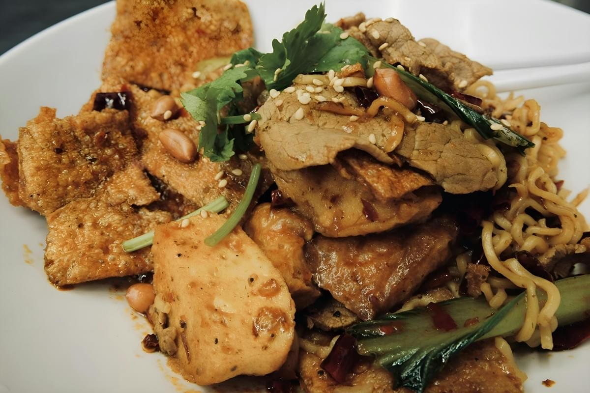 A close-up shot of a savory noodle dish served in a white bowl, featuring stir-fried noodles topped with thin slices of beef, crispy tofu sheets, and fresh bok choy. The dish is garnished with whole peanuts, sesame seeds, and cilantro sprigs.