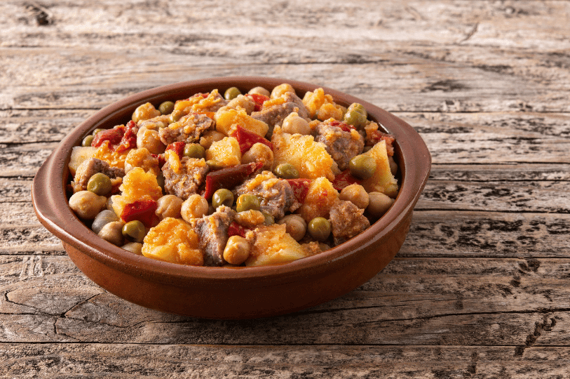 A rustic clay bowl filled with a hearty beef stew containing tender meat chunks, chickpeas, green peas, and diced potatoes in a light tomato broth, set on a weathered wooden table.