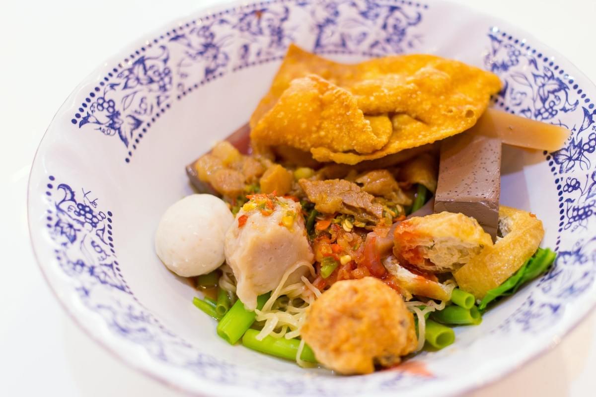  This bowl of Rojak features a colorful array of ingredients including thin noodles, fish balls, fried tofu, and leafy green morning glory, all accented by a signature pink bean curd sauce. The dish is served in a blue-patterned white bowl and topped with a large, crispy fried wonton skin for added texture.