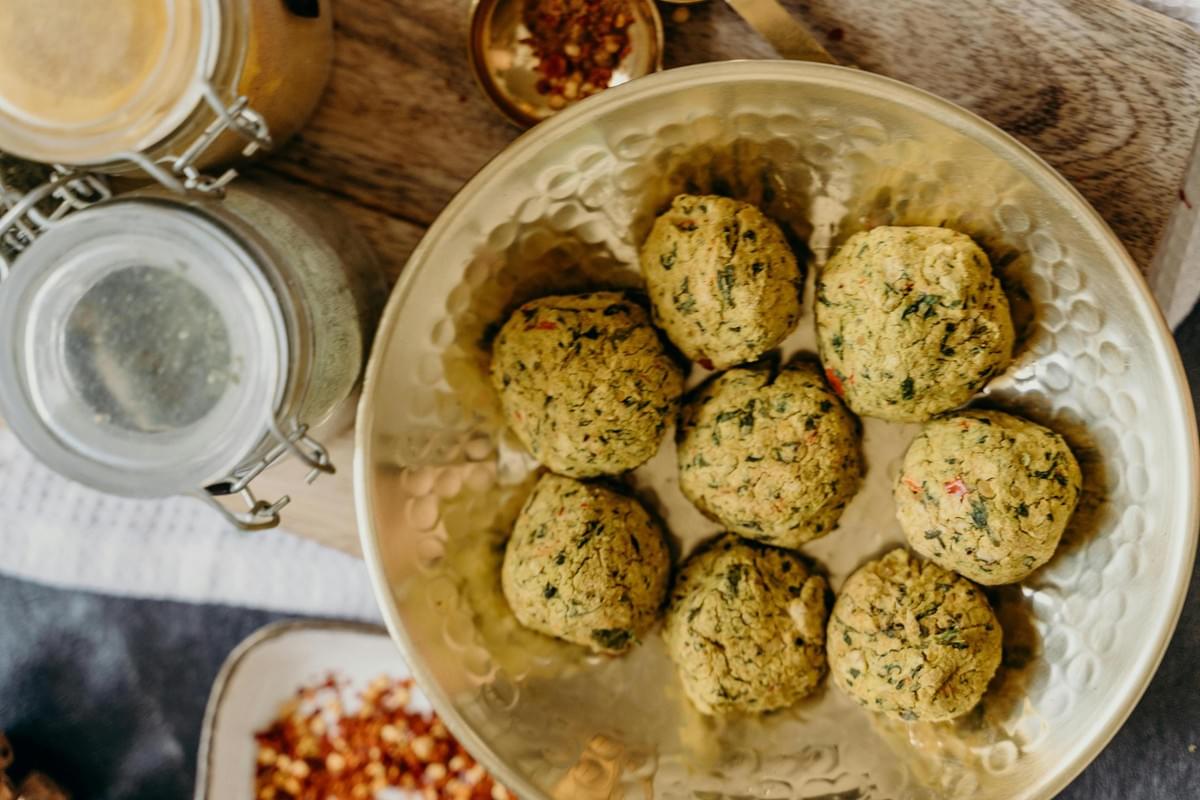 A hammered gold bowl is filled with round, herb-flecked falafel balls, sitting prominently on a wooden board. Nearby, glass spice jars and small dishes of crushed red pepper flakes add vibrant color and texture to the culinary setup.