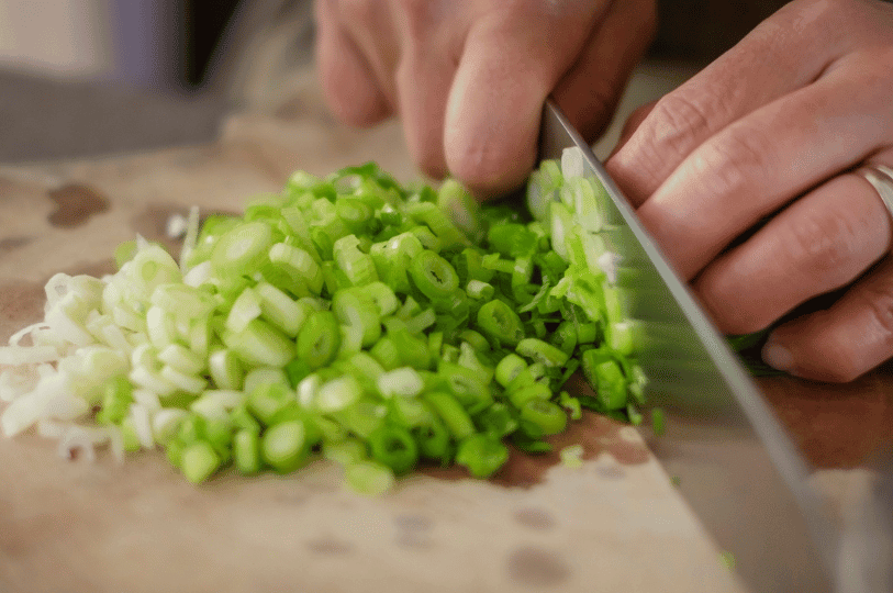 A chef’s hands using a silver knife to finely chop vibrant green leek stalks on a wooden cutting board.