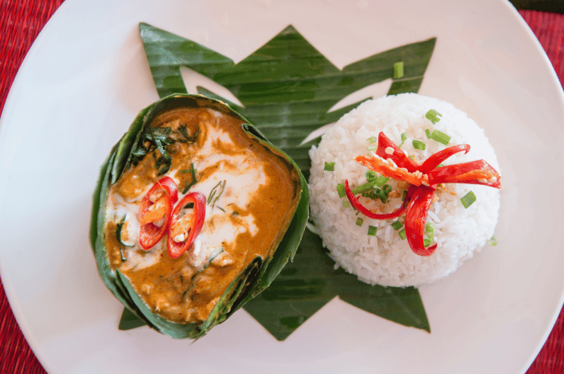 A side-angle shot of Cambodian steamed fish curry presented in a deep banana leaf cup, topped with a delicate drizzle of coconut cream, fresh herbs, and red chili, resting on a wooden tray.
