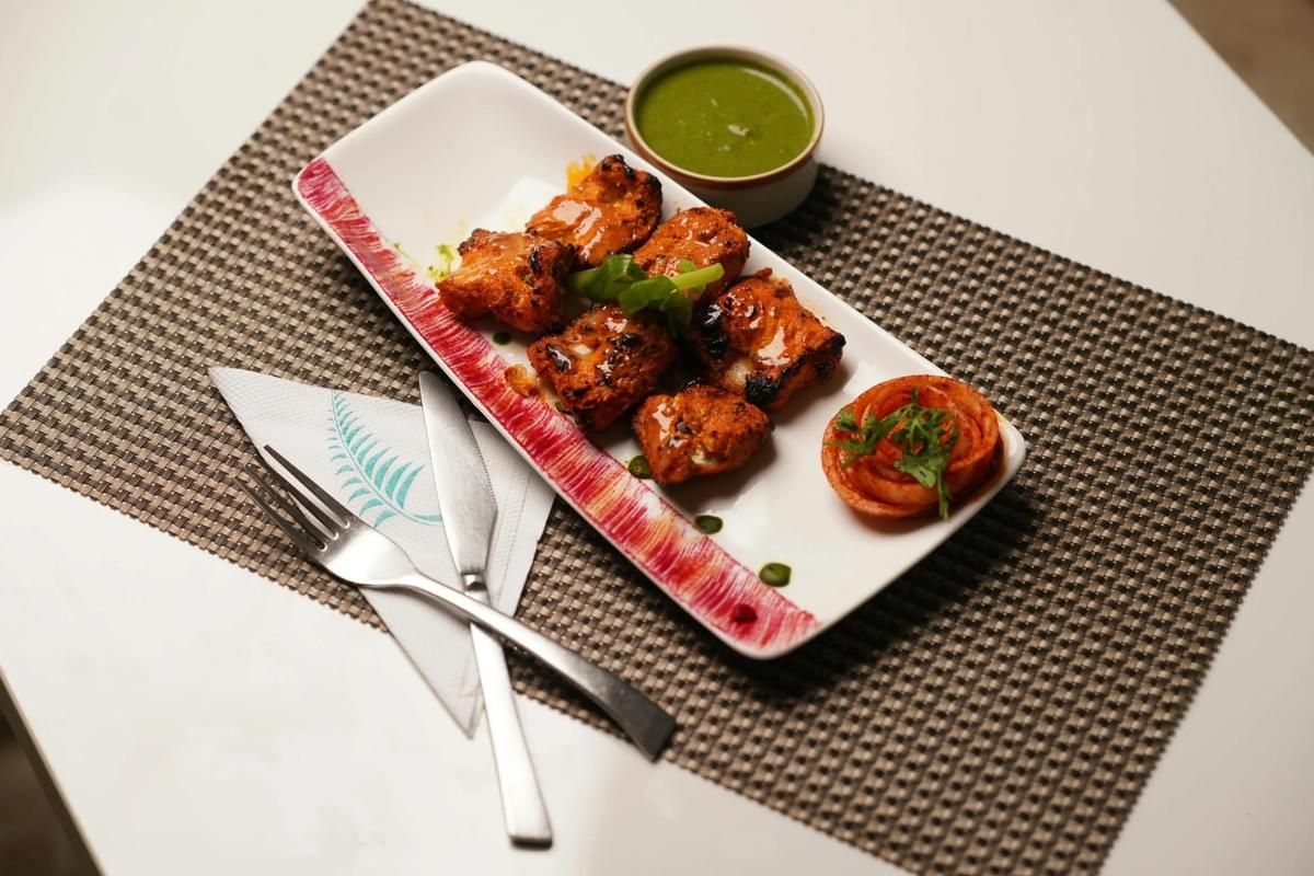 Pieces of marinated, grilled chicken are served on a white platter alongside a small bowl of green chutney and a garnish of sliced onions. The dish is presented on a textured placetmat with a fork and knife resting on a white napkin.