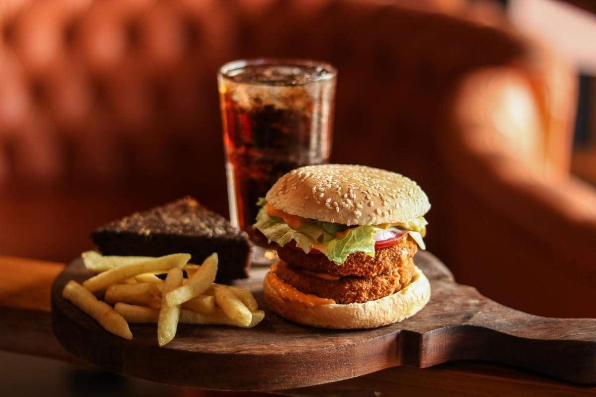 A double-patty crispy burger topped with lettuce, tomato, onion, and sauce is served on a wooden board alongside golden French fries and a slice of chocolate cake. In the background sits a tall glass filled with a cold soda and ice, set against a warm, softly blurred backdrop.