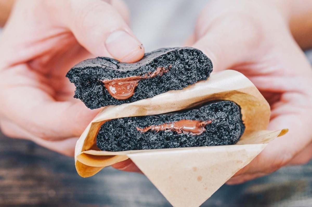 Two hands hold open two halves of a dark charcoal cookie to reveal a gooey, melted milk chocolate filling. The bottom half rests inside a piece of light brown parchment paper.