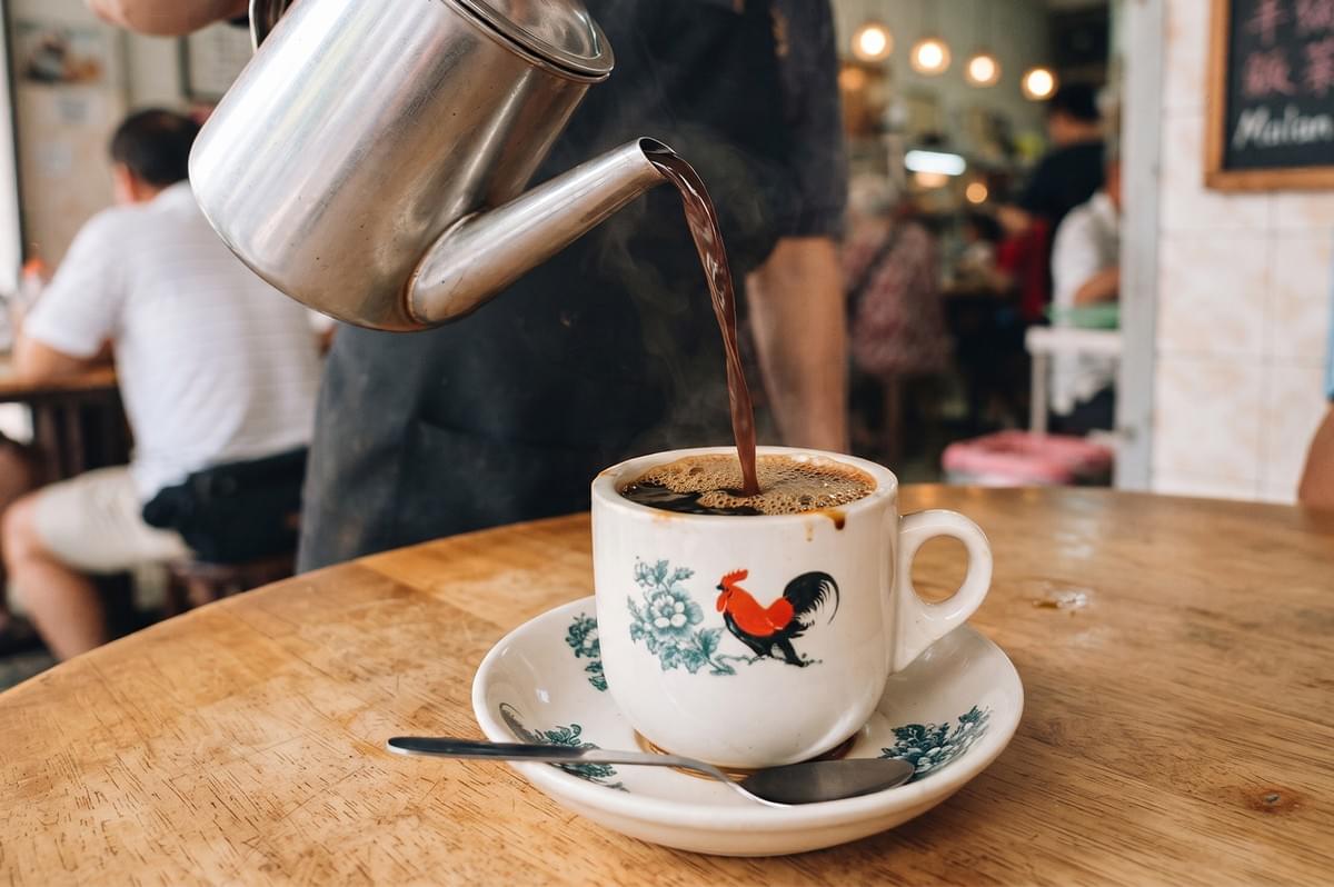 Traditional Singaporean kopitiam coffee pouring into a classic rooster cup at a local hawker stall.